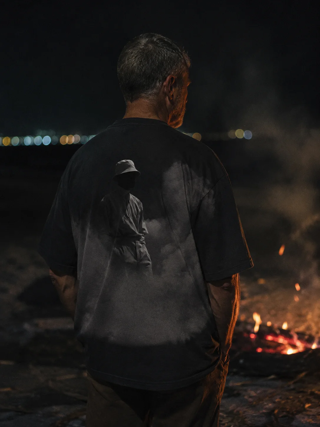 EMBER tee shot from behind on a dark beach beside a fire, a faint figure printed across the back