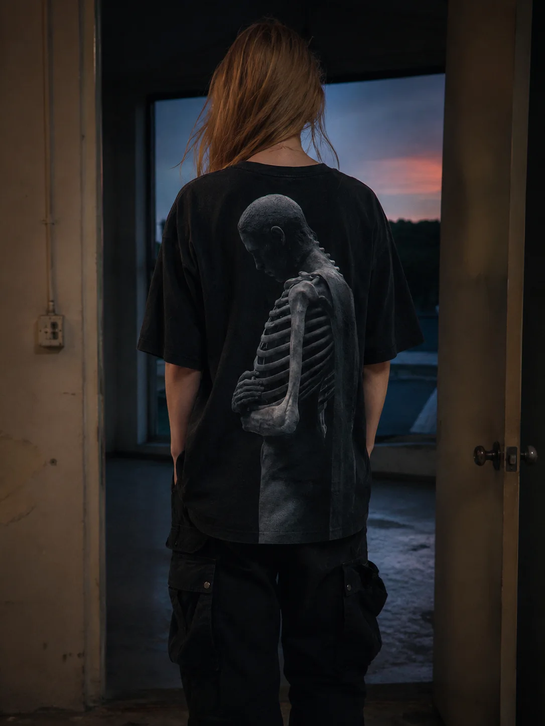 HOLLOW tee worn in a doorway at dusk, a gaunt skeletal figure printed across the back