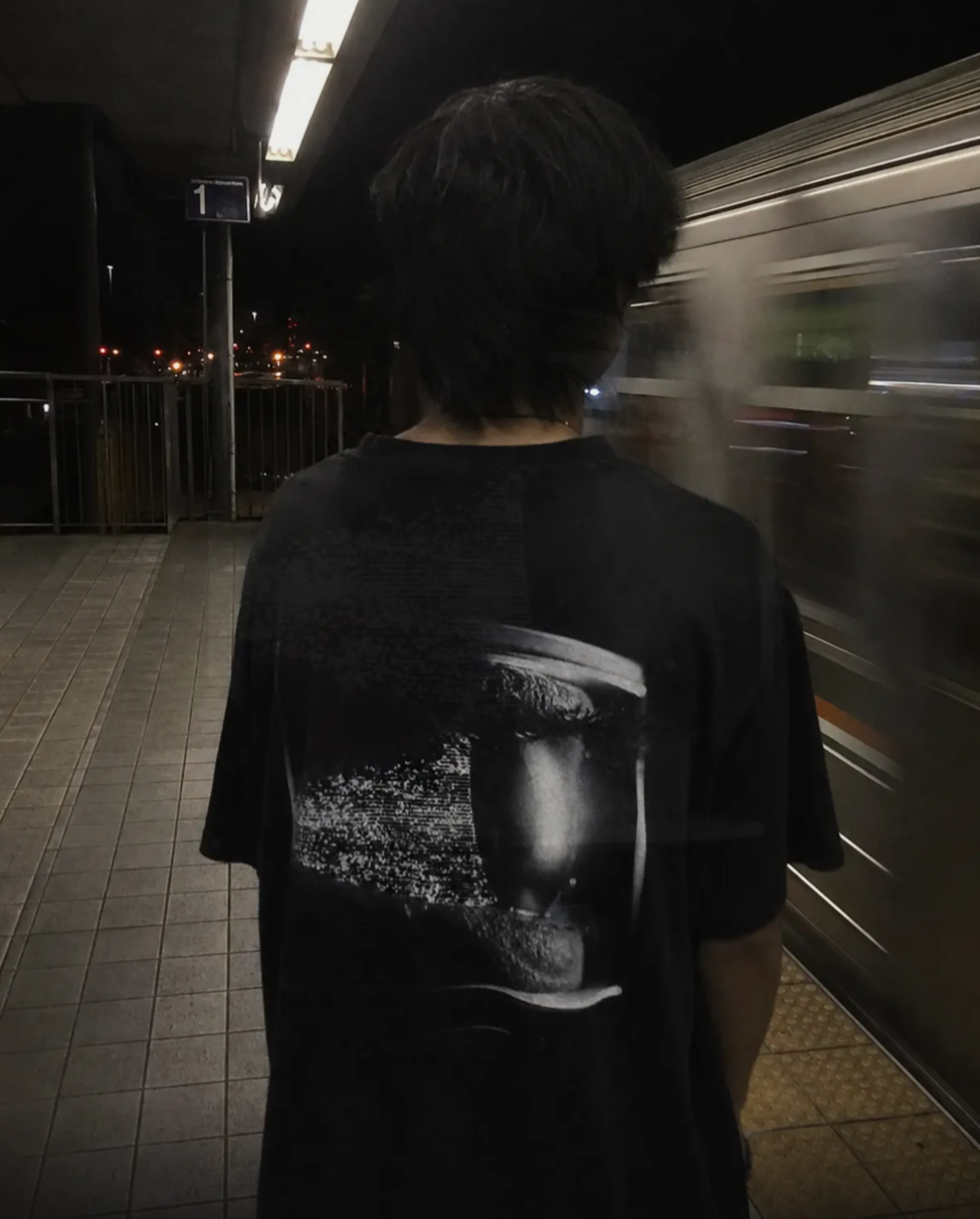 A tee worn on a night train platform, a glitched face print catching the light as a train passes