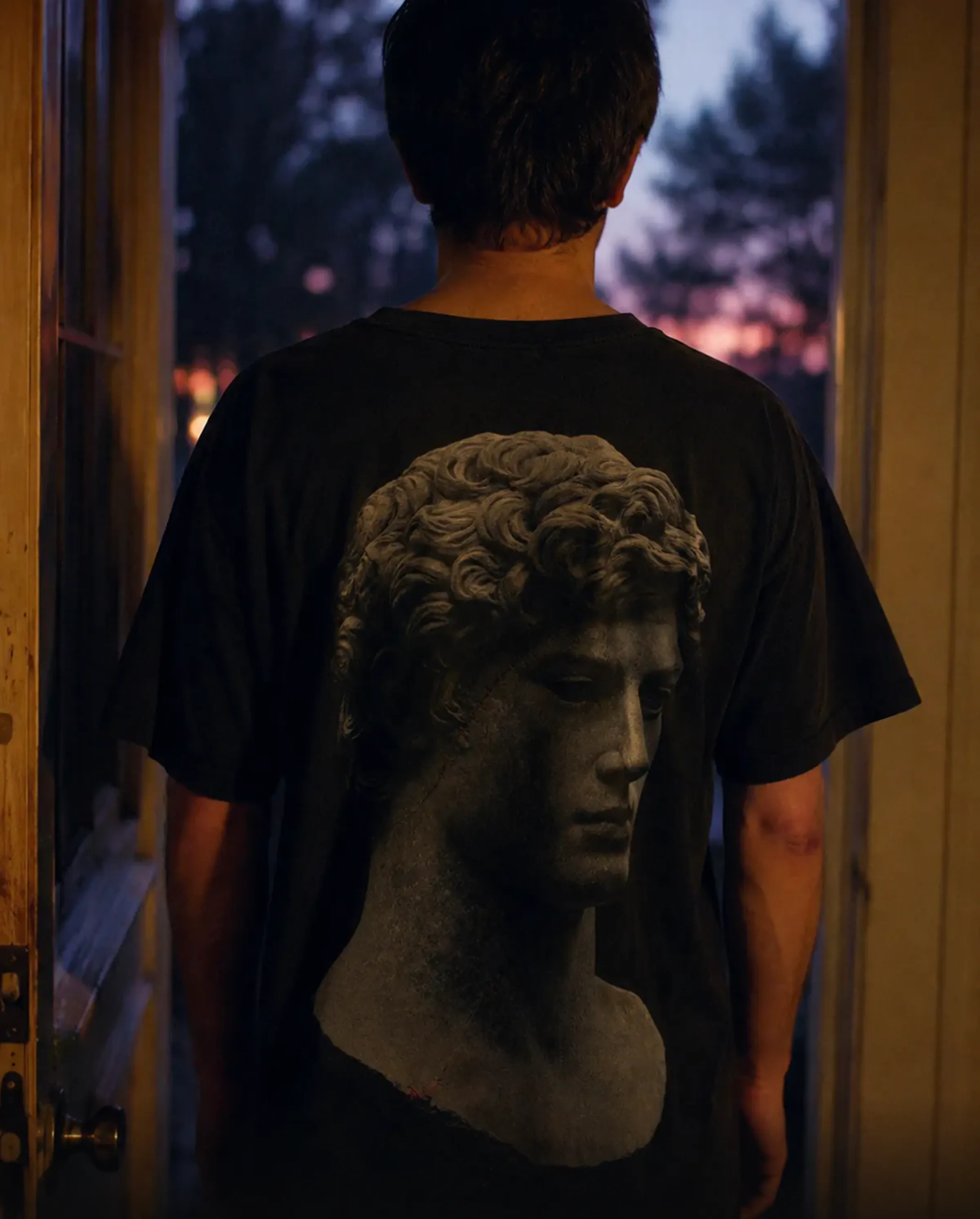 A black tee worn in a doorway at dusk, a classical marble bust printed across the back